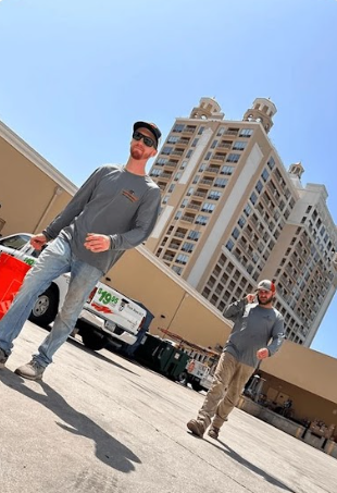 Two people walking across an outdoor concrete lot in front of a tall, beige high-rise building under a clear blue sky.