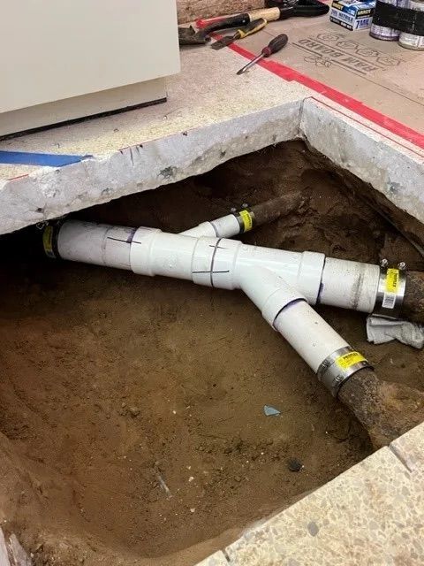 White PVC pipes connected with Y-fittings to existing pipes in a hole dug into a dirt floor.