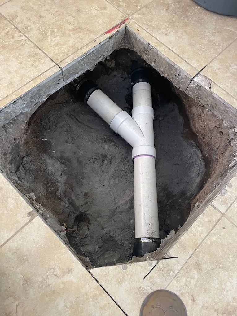 White PVC Y-pipe plumbing connector installed in a square hole cut through tiled flooring into the dirt below.