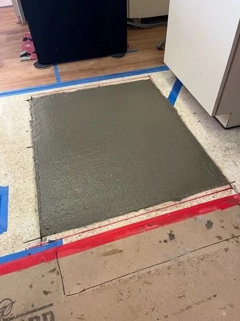 A square patch of wet concrete fills a rectangular opening in an indoor floor, framed by blue and red painter's tape.