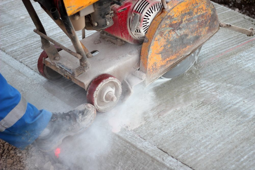 A person in blue work pants uses a walk-behind saw to cut a concrete surface, creating a plume of dust.