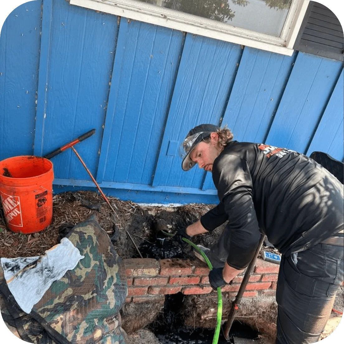 A person in a black shirt and cap works on a repair in a dirt trench next to a blue wall, using a green hose and a tool.