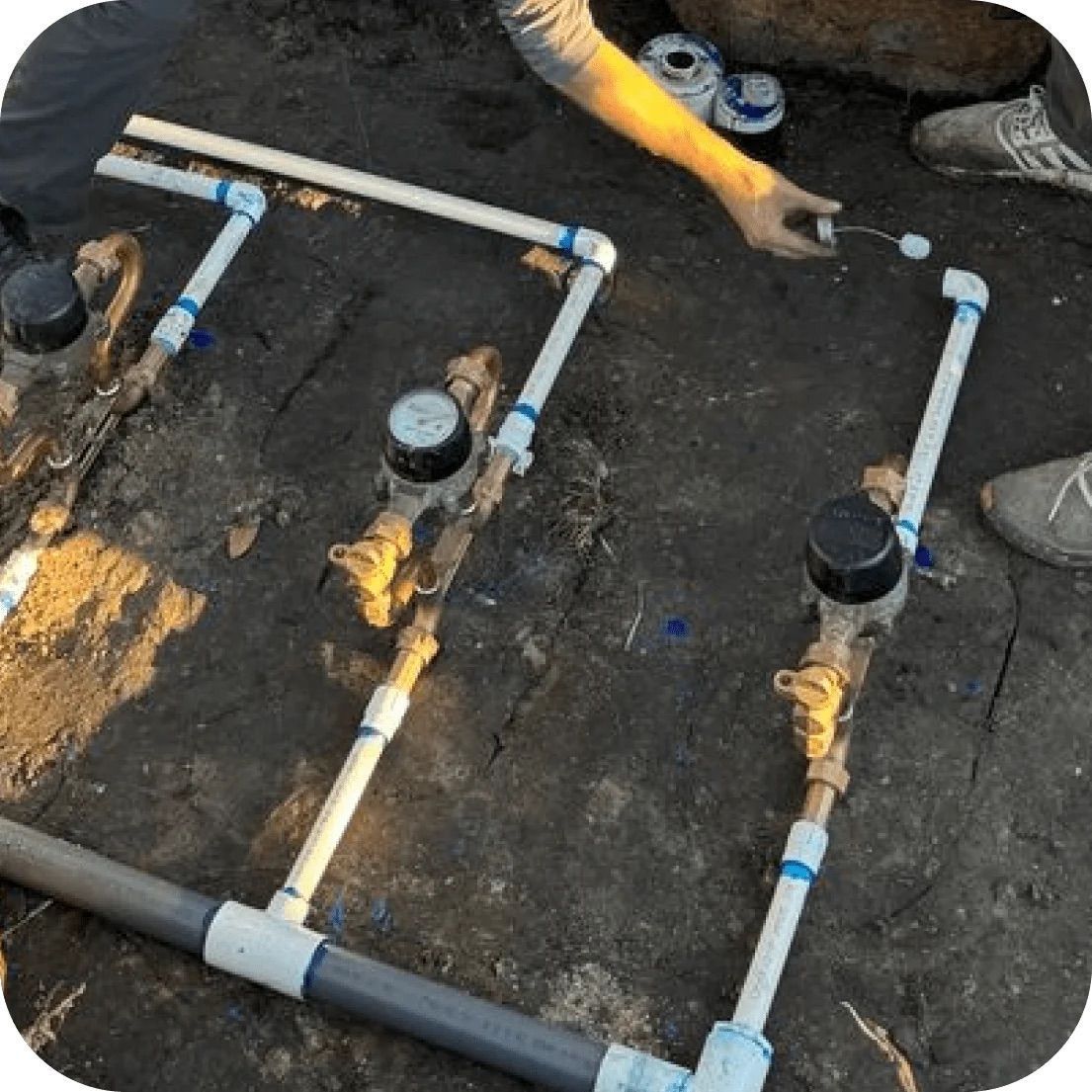A worker repairs white PVC plumbing pipes connected to water meters embedded in the dirt ground.