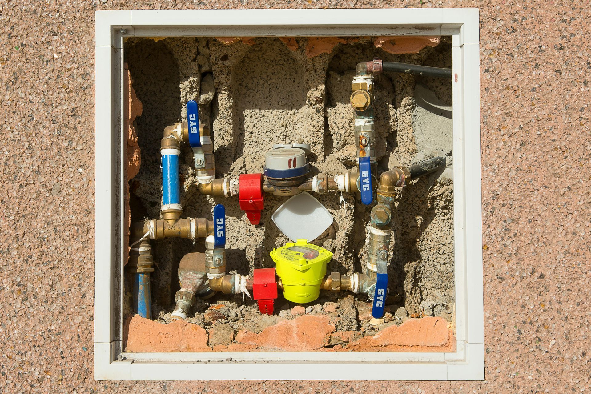 Water meter and pipes within a recessed wall enclosure. Red, blue, and yellow valve handles.