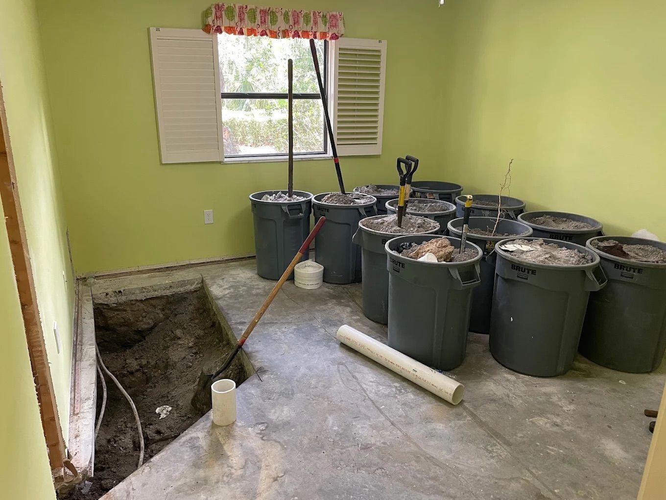 Room undergoing renovation with debris bins, exposed concrete floor, and hole. Green walls and window.