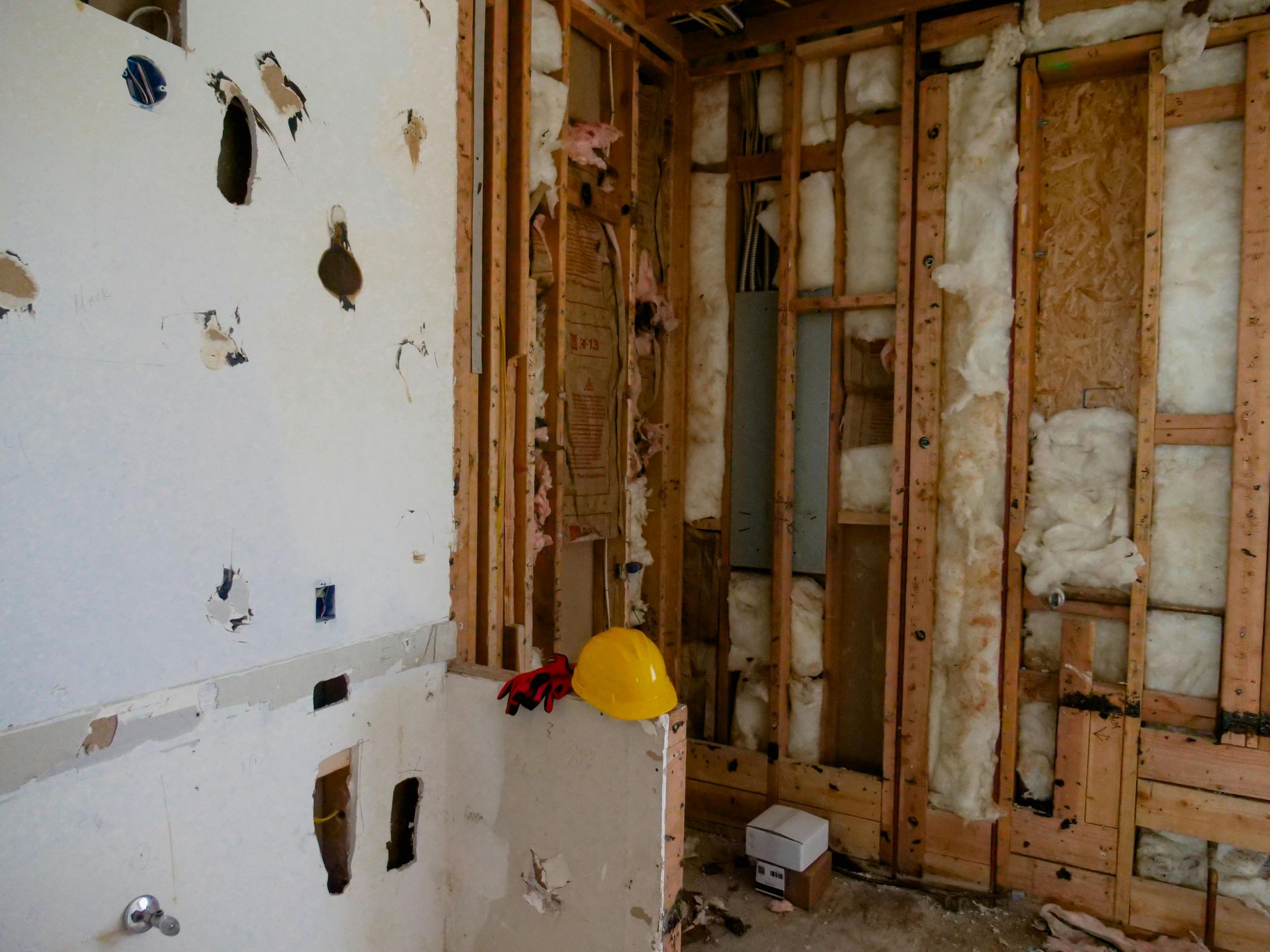 Interior wall under construction with exposed studs, insulation, and damaged drywall. A yellow hard hat rests on a ledge.