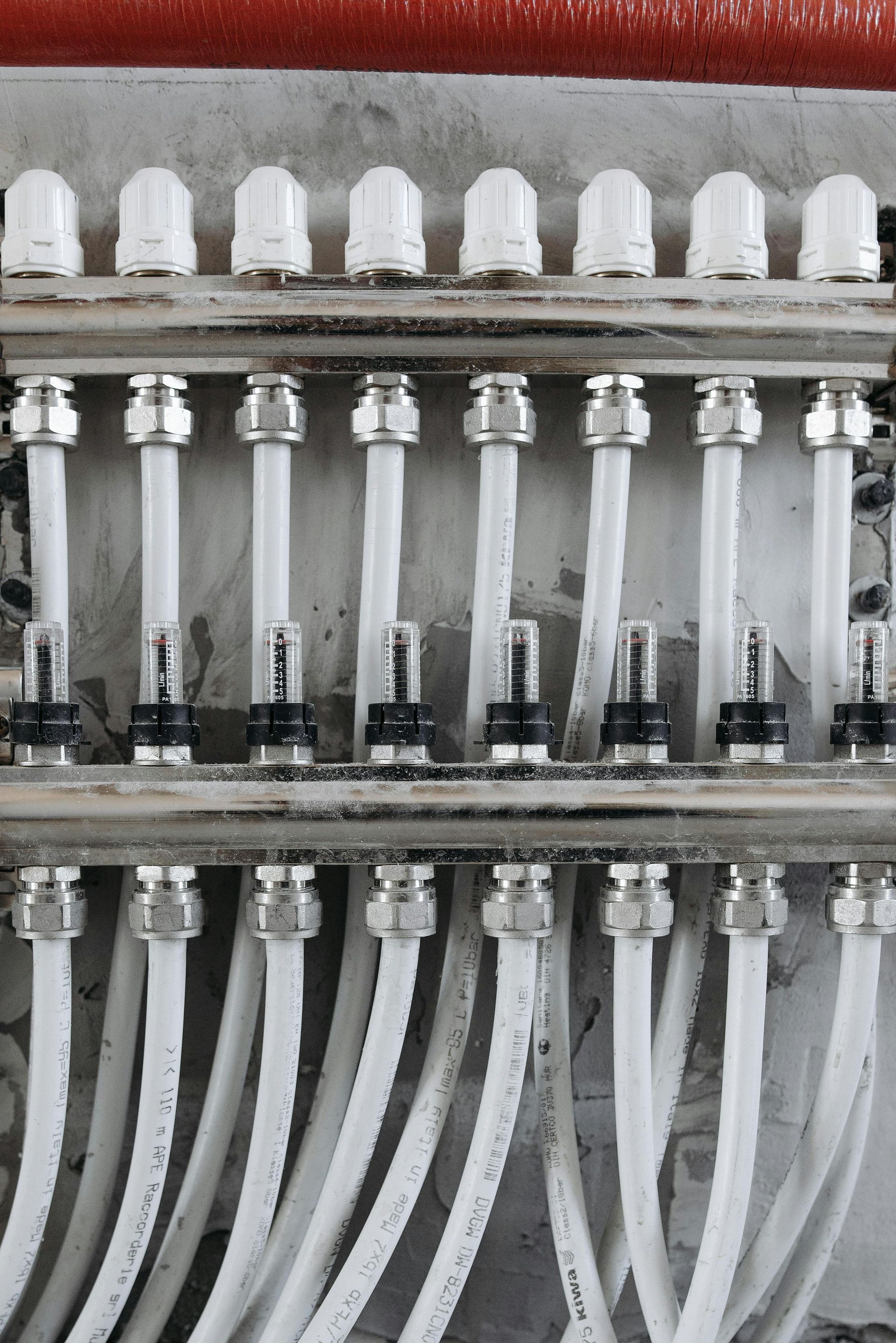 White pipes and connectors in a radiant floor heating manifold system, mounted on a concrete wall.