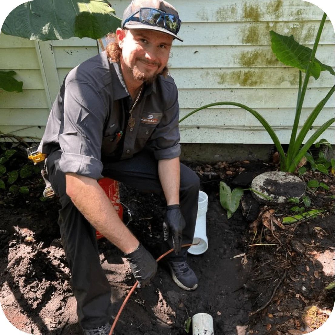 Plumber kneeling near pipe, using a tool. Outdoors near a building.