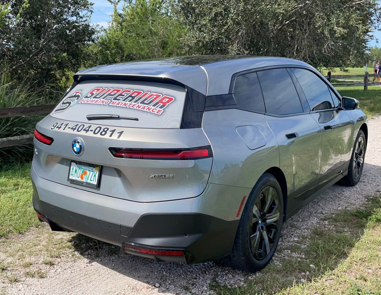 Gray BMW iX SUV parked on a gravel path with Superior Auto Glass advertisement on the rear window.