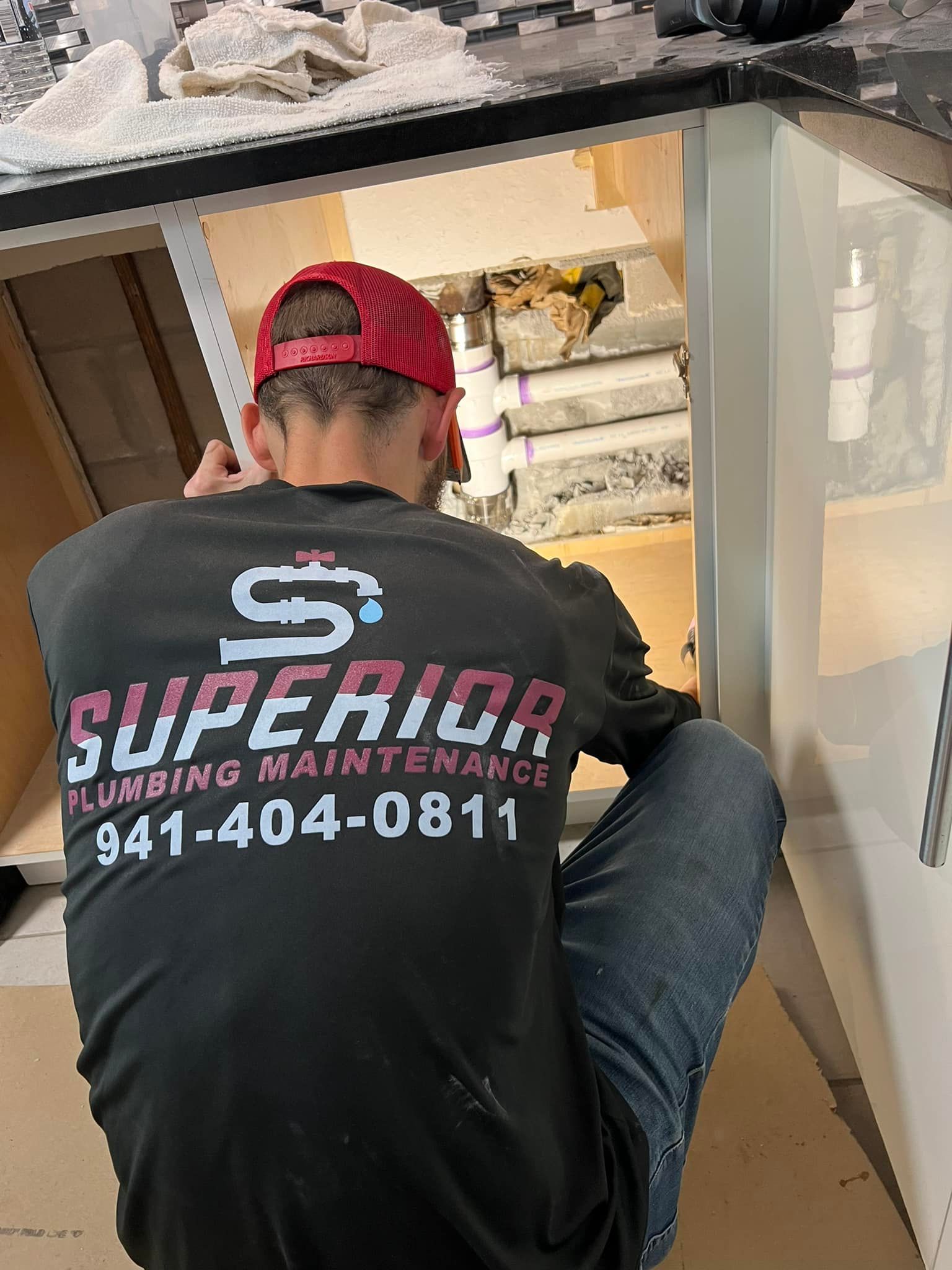 Plumber working under a sink. Wearing a red hat, a shirt that says 