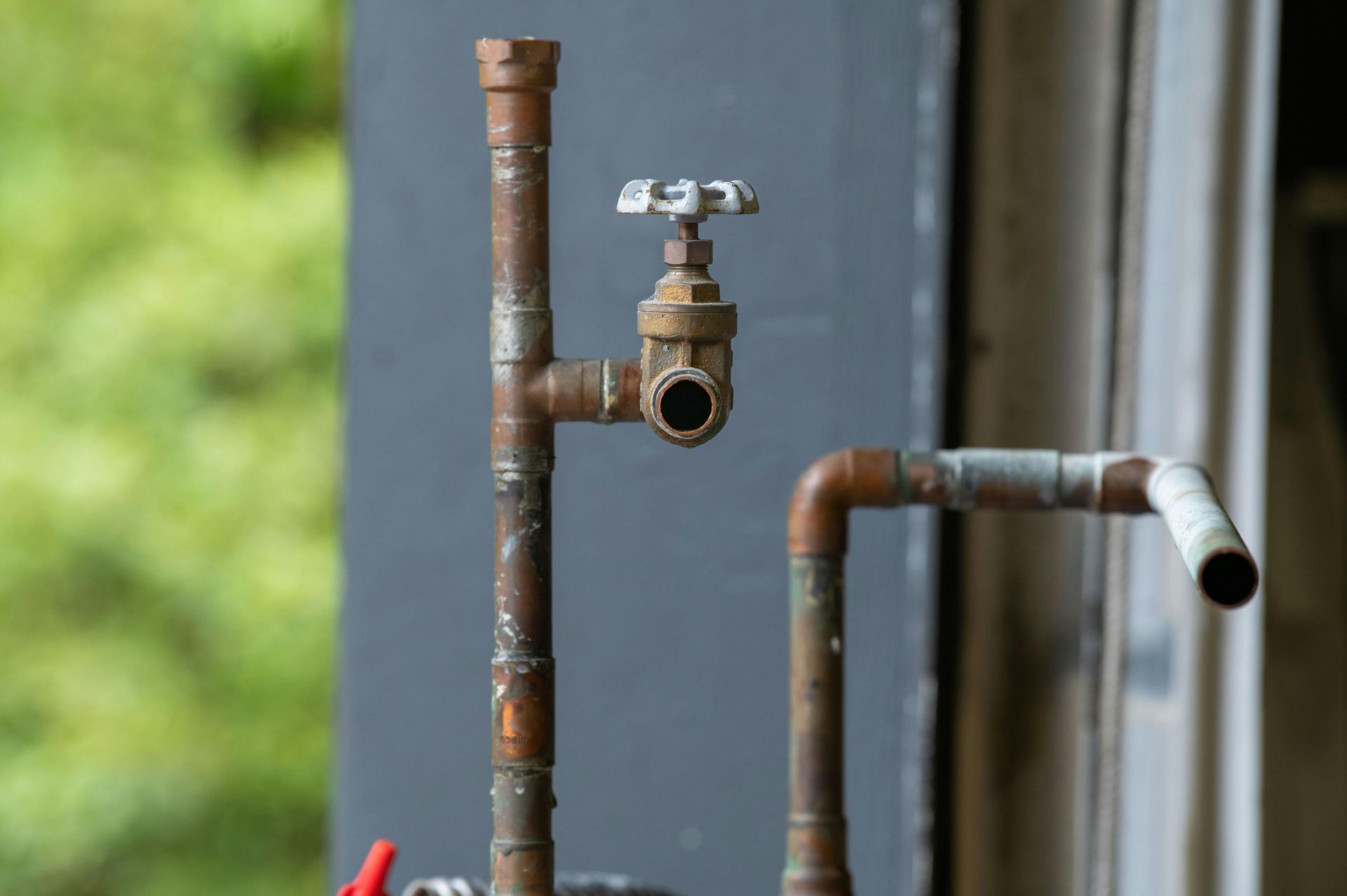 Rusty copper plumbing with a valve and pipes against a blurry green and gray background.