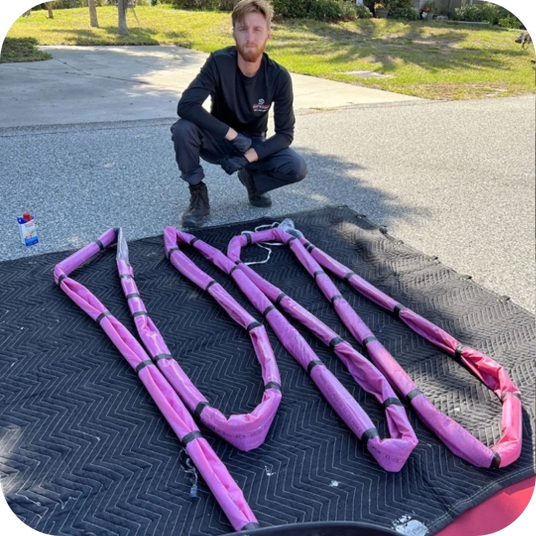 Man squats near folded pink lifting slings on a mat in a driveway.