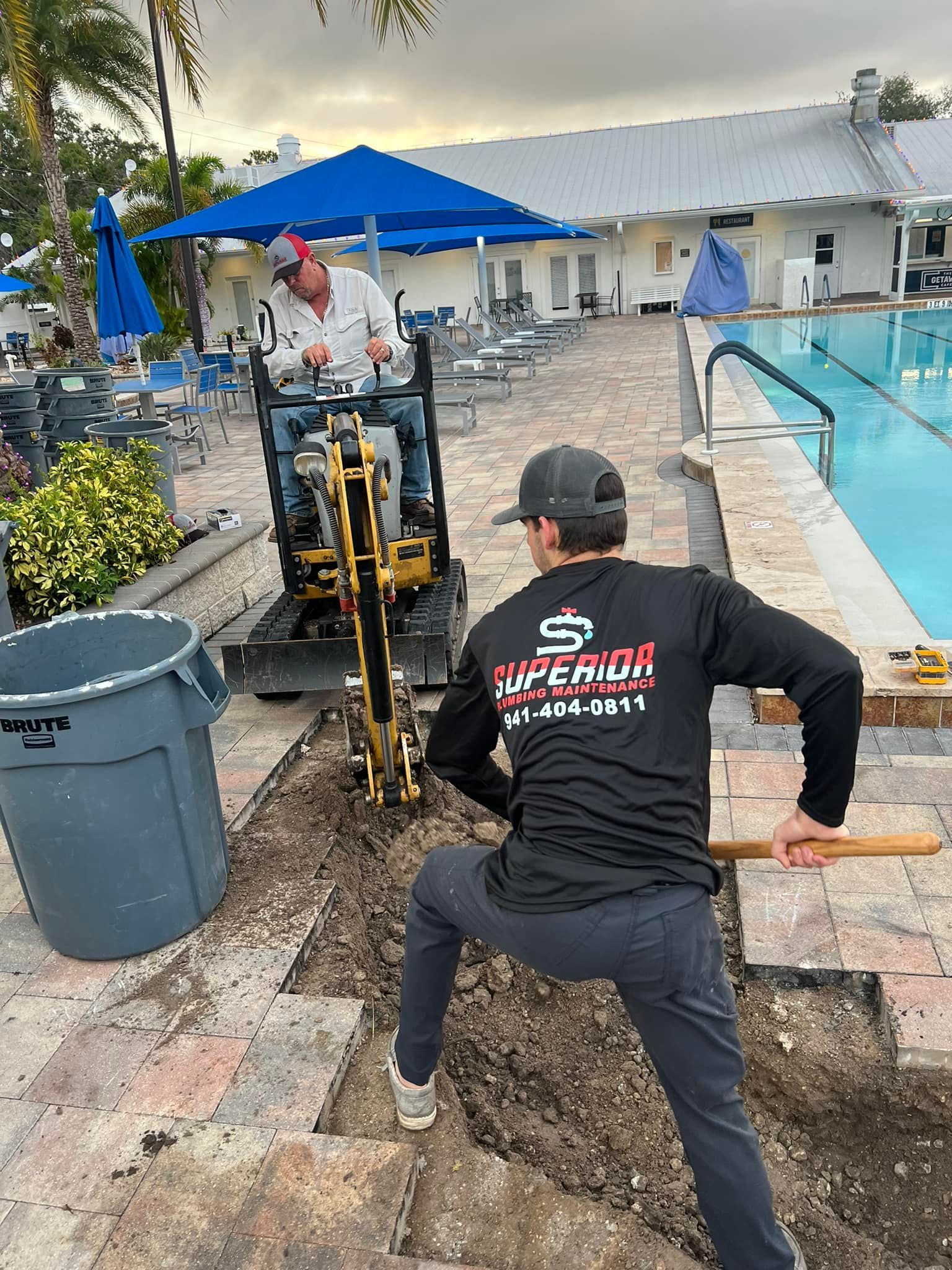 Two workers use a small excavator and shovel to dig near a pool. One worker operates the machine; the other uses a shovel.