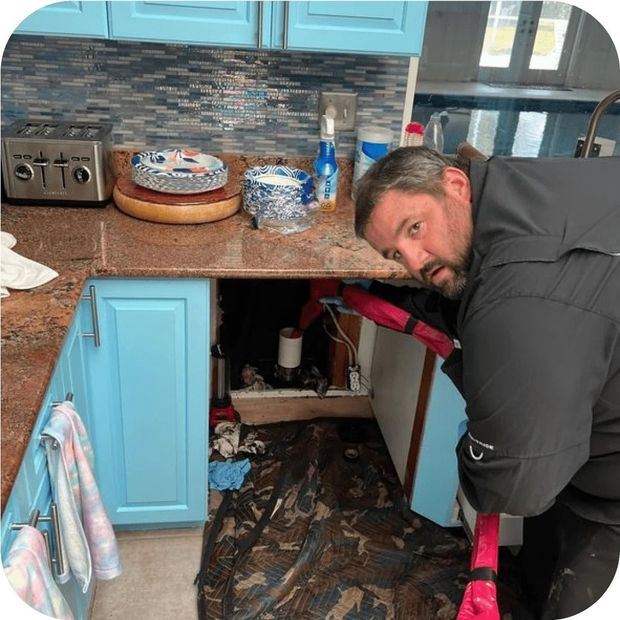 Man peering under a kitchen sink. Kitchen has blue cabinets, brown countertop, and a water leak.