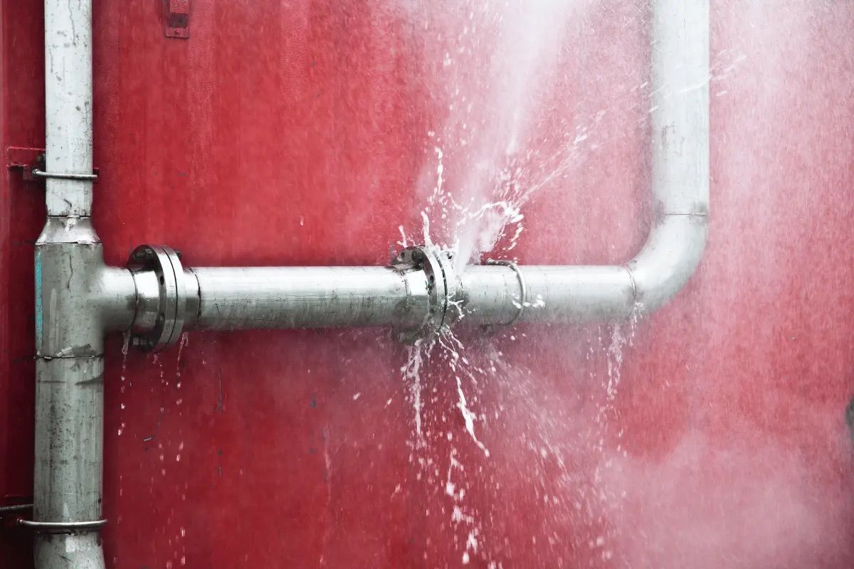 A metal pipe, connected by a joint, bursts with water spraying on a red wall.