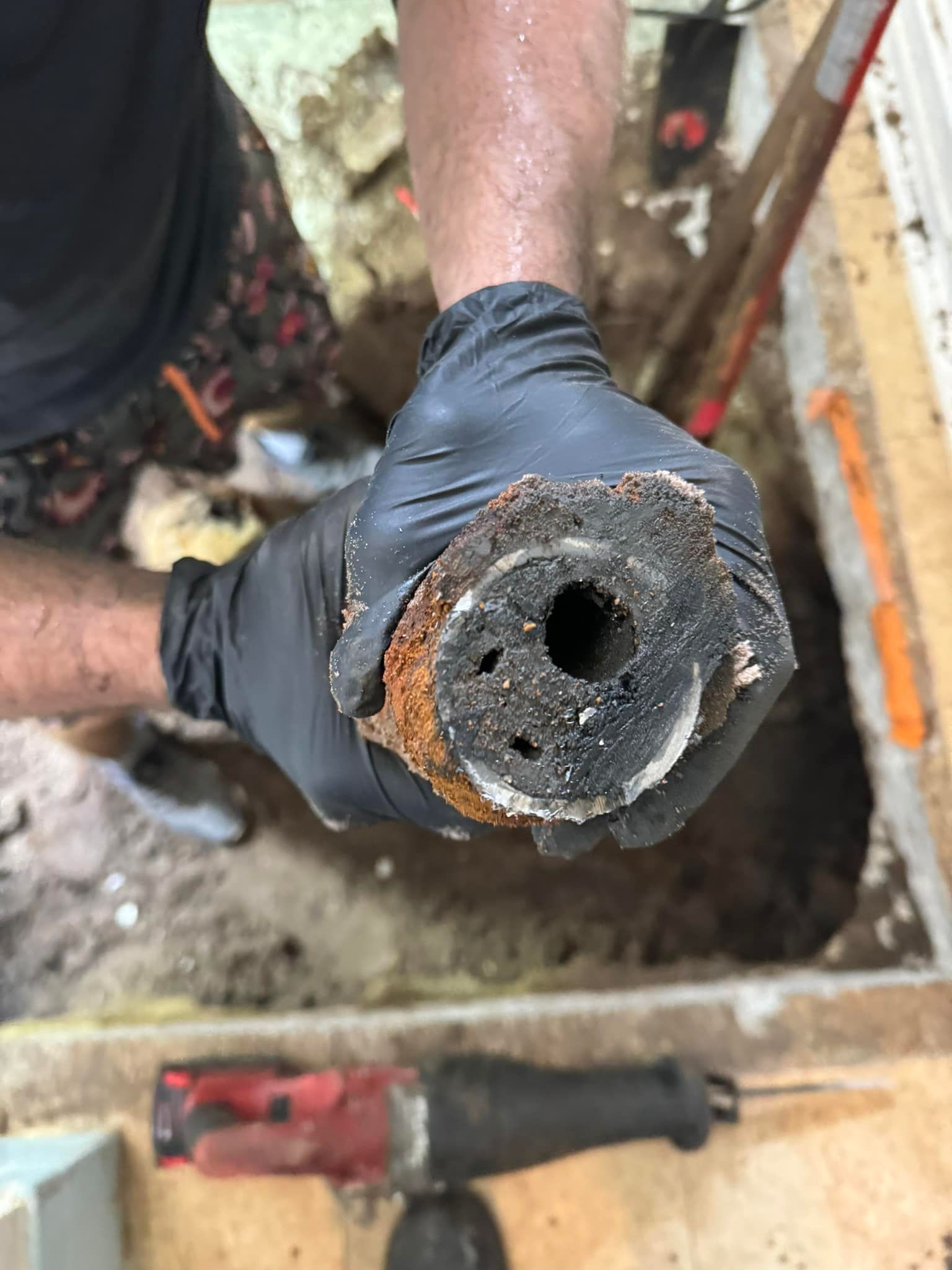 A person in black gloves holds a corroded pipe segment, near a hole in the ground with a saw below.
