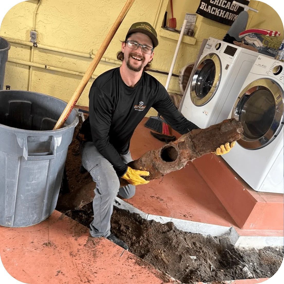 Man holding large rusty pipe in a basement. Smiling, wearing a hat, gloves, and work clothes.