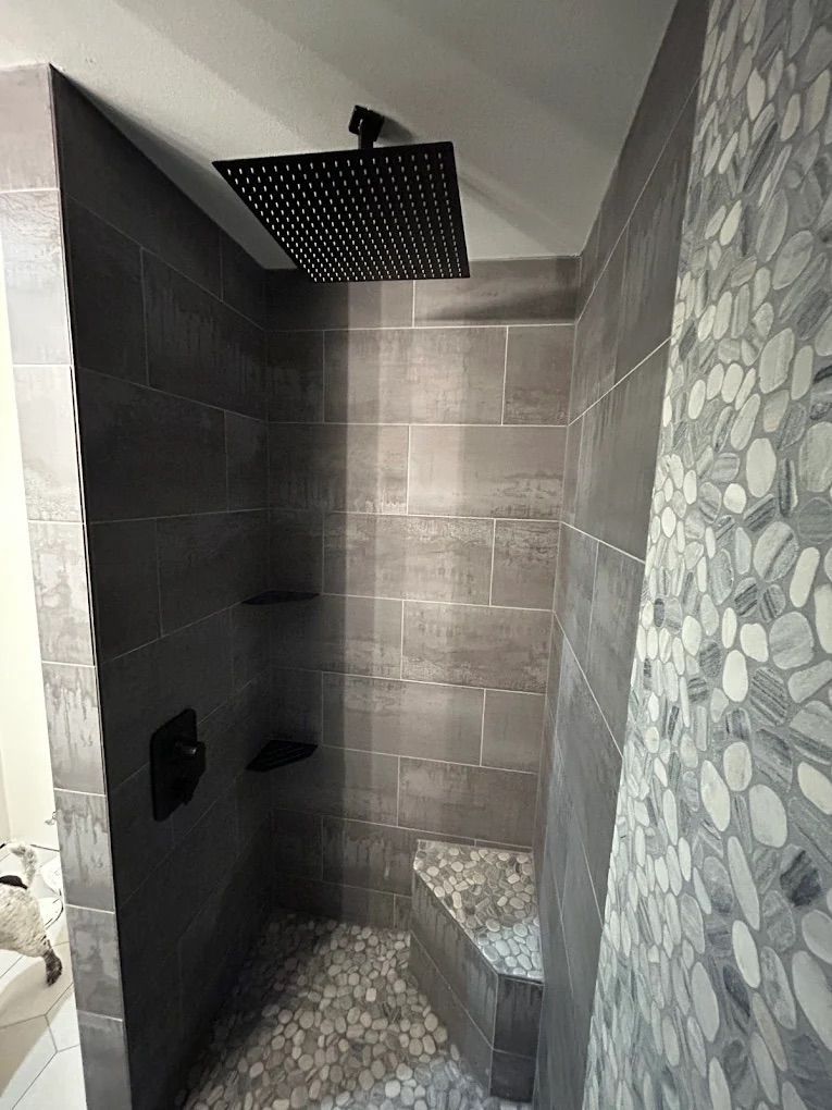 A modern shower with dark gray tile walls, pebble floor, and a black square rain showerhead.