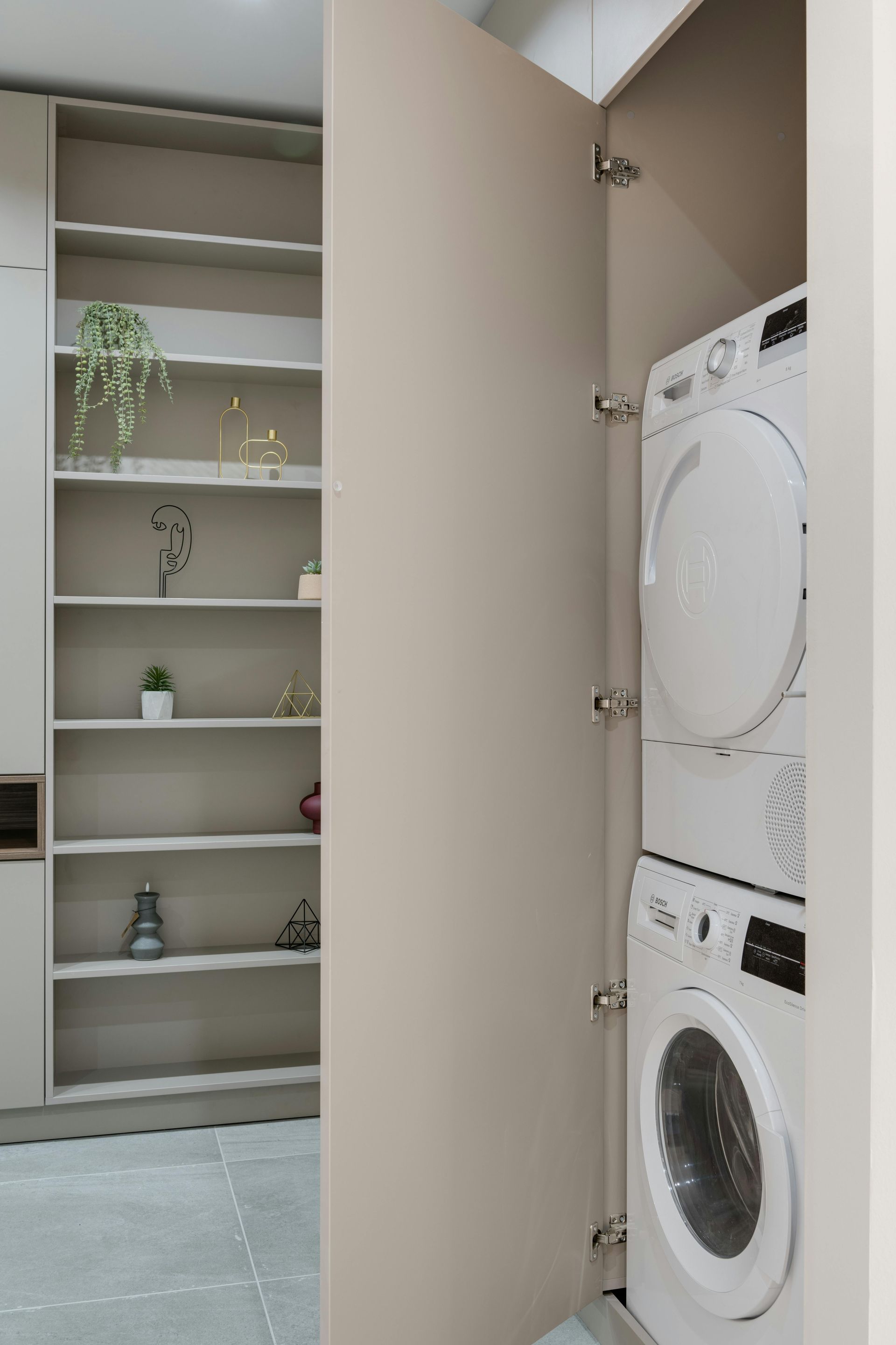 Built-in laundry closet with a stacked washer and dryer, next to shelving, all in neutral tones.