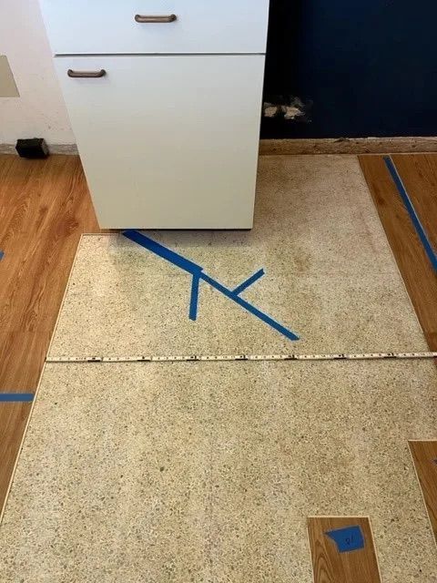 White cabinet on speckled flooring, blue tape markings, measuring tape, and wood flooring border.
