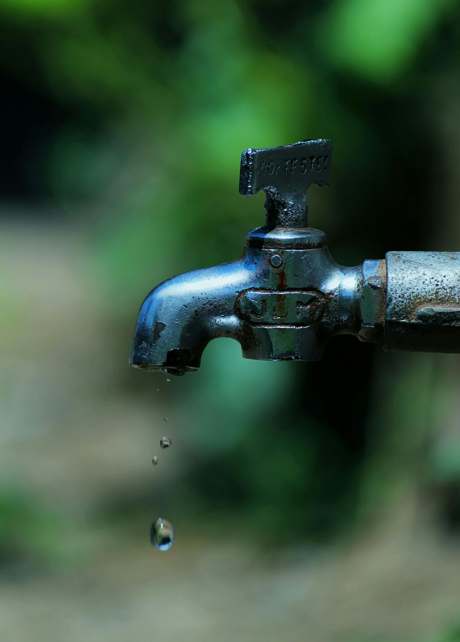 Dark metal outdoor faucet dripping water. Green, blurred background.
