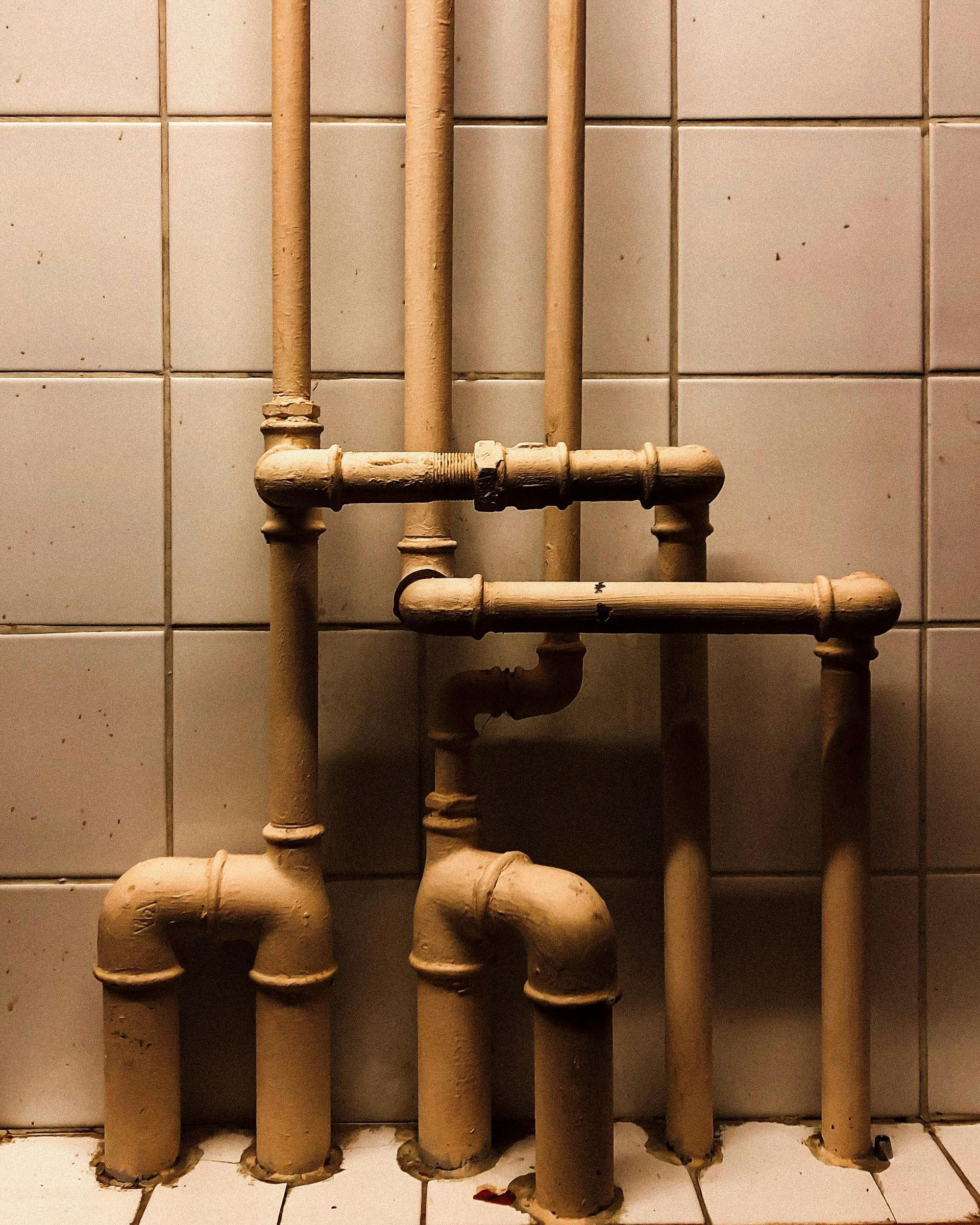 Pipes against tiled wall, connected by elbows. Beige color, dark setting.