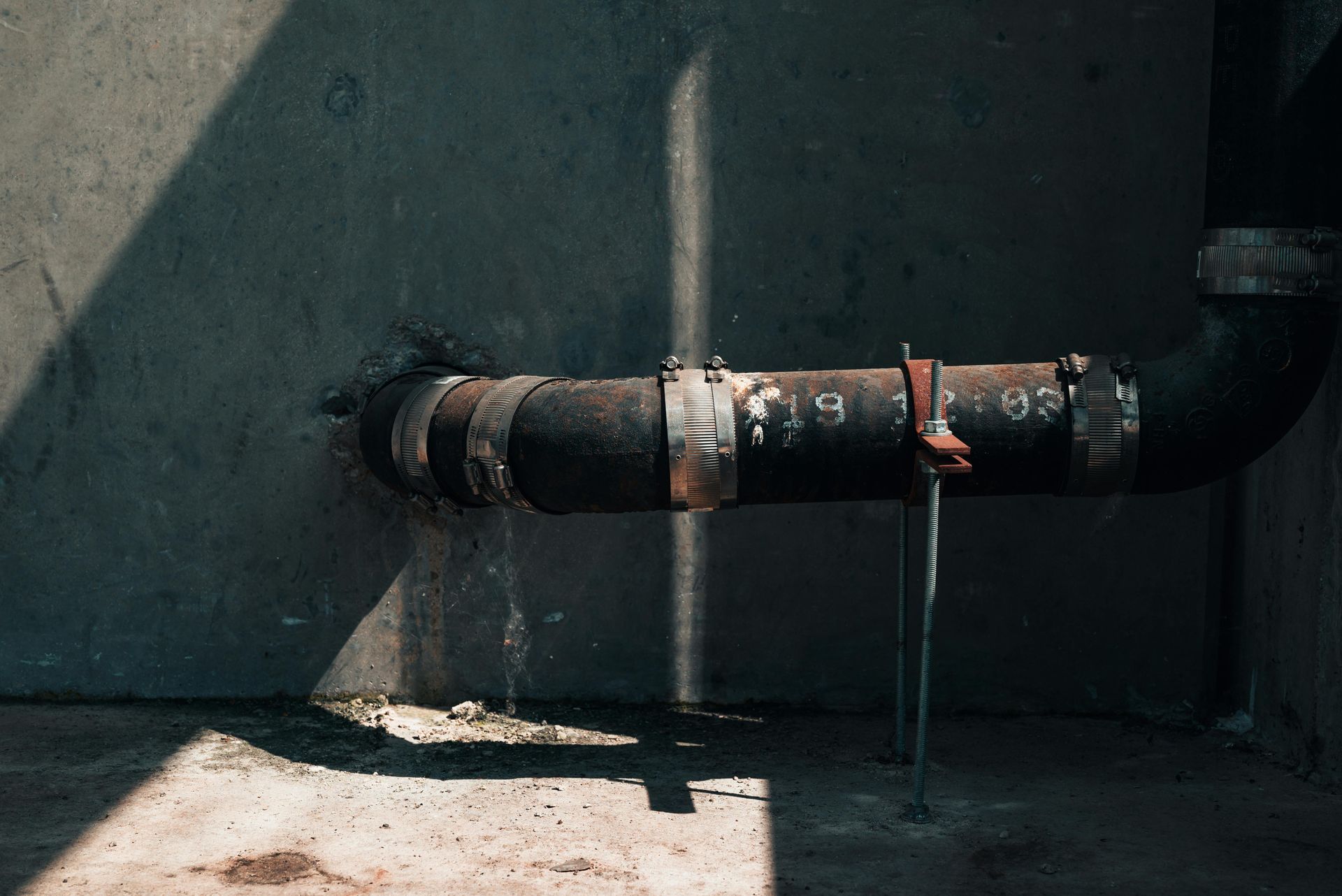 Black pipe attached to a concrete wall; sunlight creates shadow and highlights rust.
