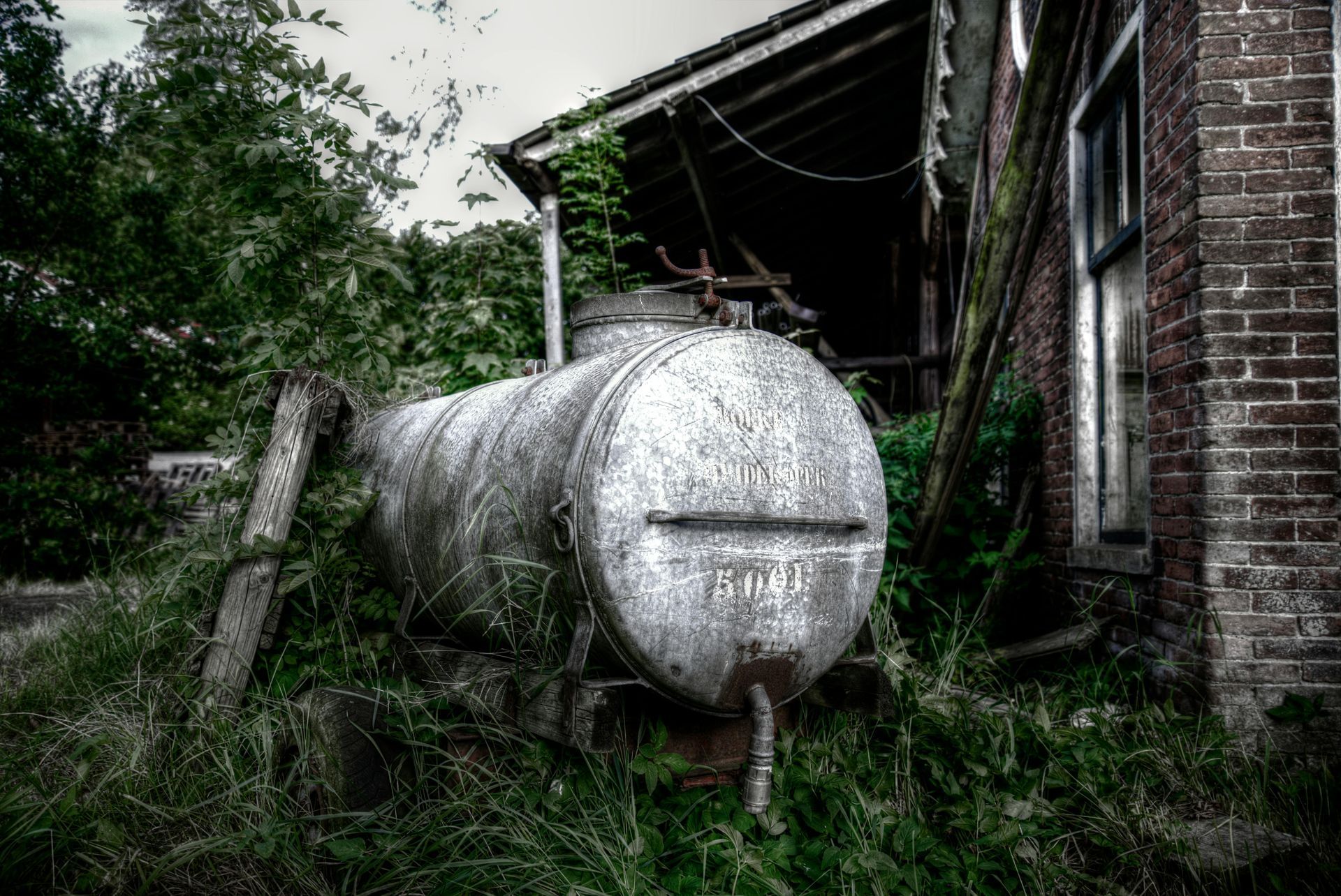 Dilapidated, rusty metal tank in overgrown grass, next to a crumbling brick building.