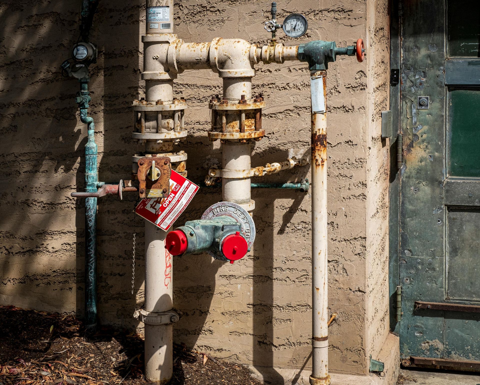 Pipes against a textured wall with gauges, valves, and a red fire department connection next to a weathered green door.