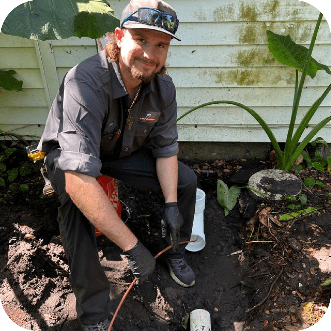 Plumber in work clothes, smiling while working with a pipe in a yard near a building.