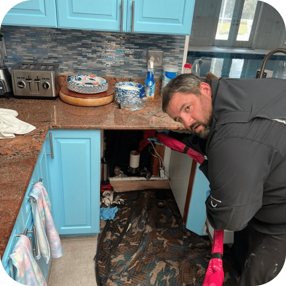 Man working under a kitchen sink with blue cabinets and a mottled backsplash.