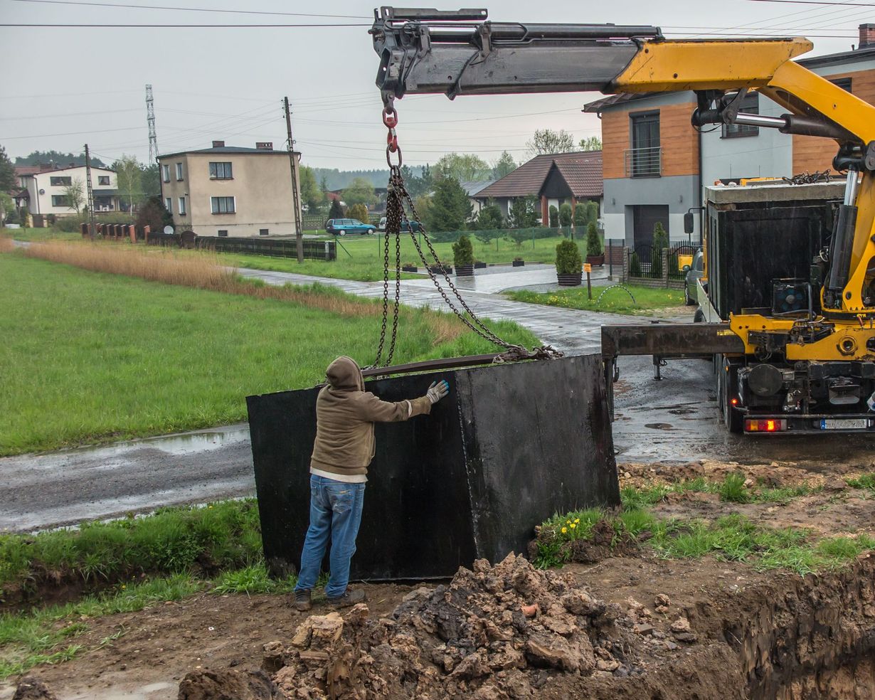 A crane lowers a large, black, rectangular septic tank while a person in work clothes steadies it on a construction site.