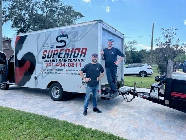Two people stand by a Superior Plumbing Maintenance truck in a paved driveway on a sunny day.