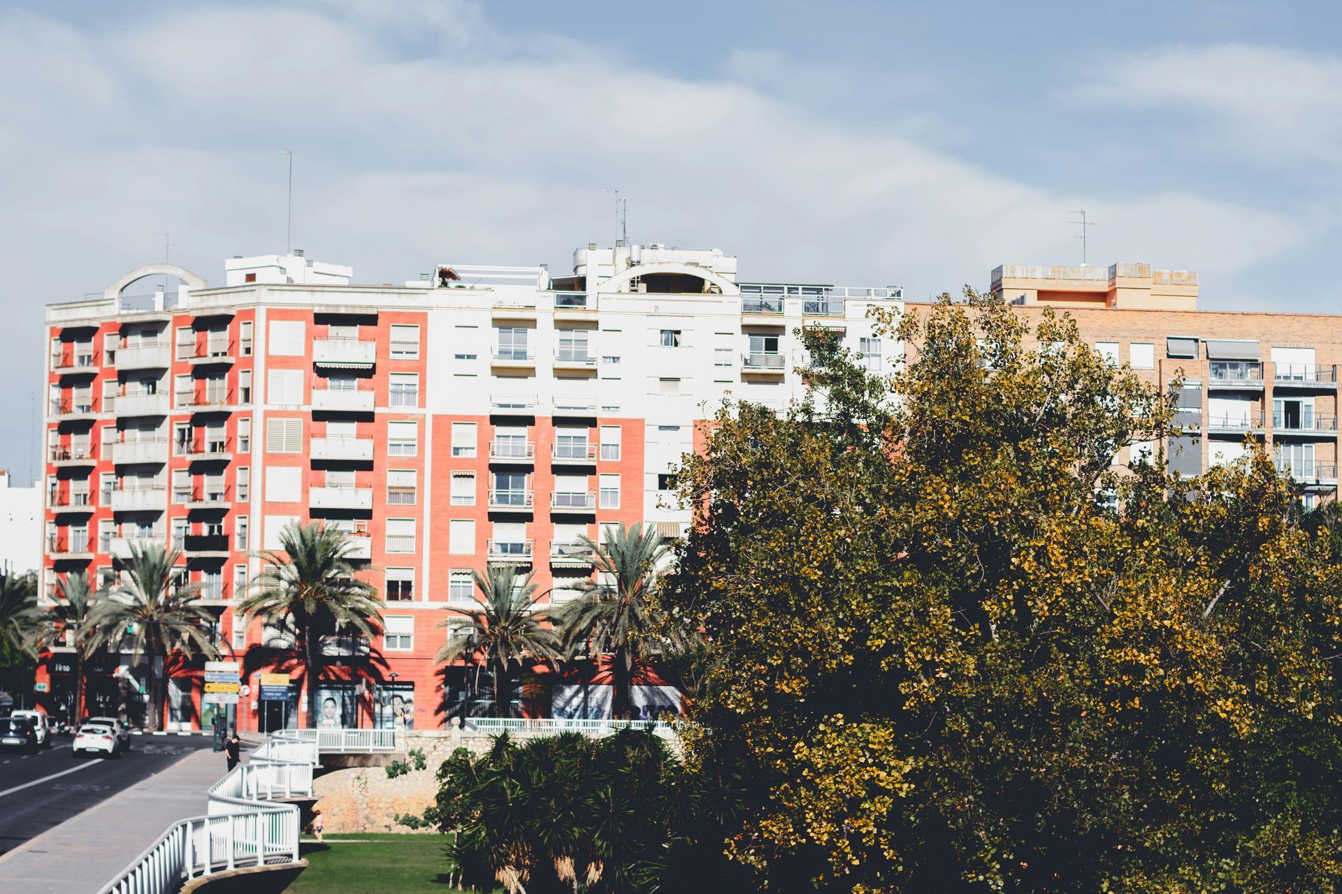 A multistory apartment building with red and white facades, surrounded by palm trees and greenery under a clear blue sky.