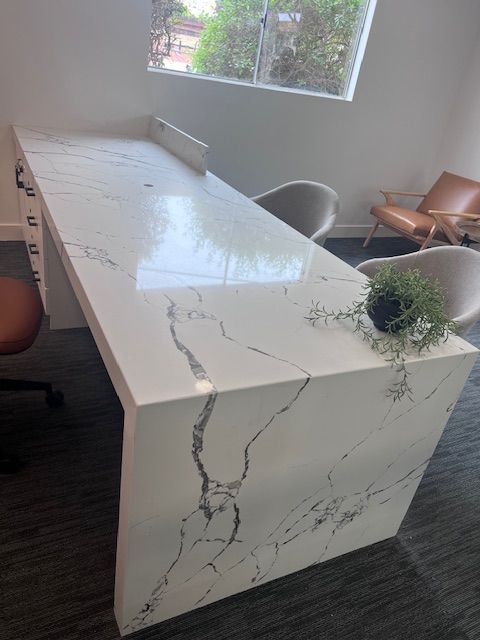 White marble countertop with black veining. Black office chairs, potted plant, and windows in background.