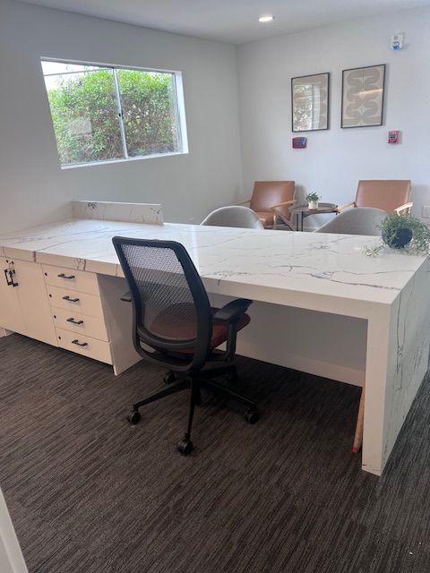 Office with large white marble desk, black mesh chair, window, and waiting area with chairs and art.