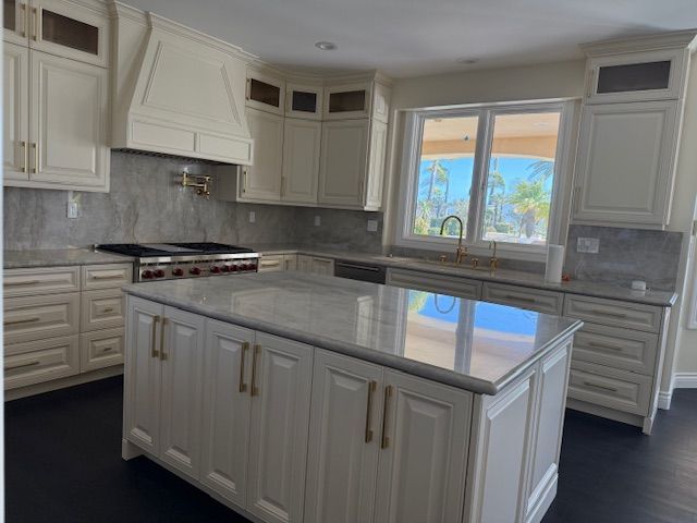 Cream-colored kitchen with island, cabinets, and range. Gray countertops, brass hardware, dark floors, and a window with view.