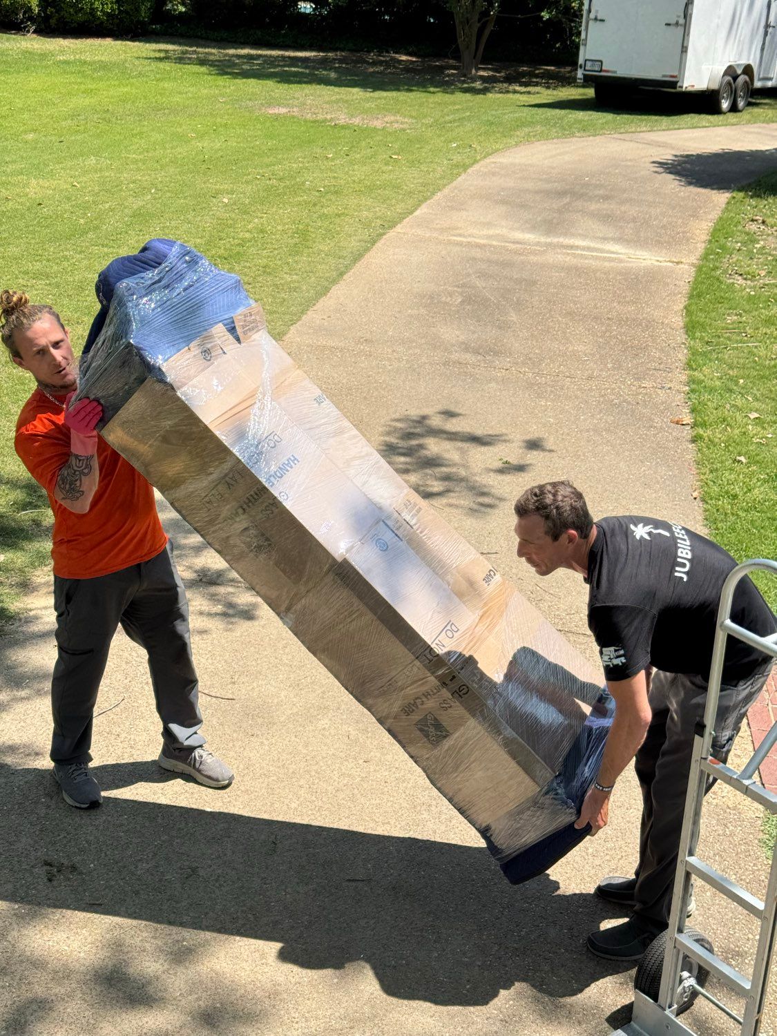 Two workers carrying a large wrapped panel on a driveway beside a grassy lawn