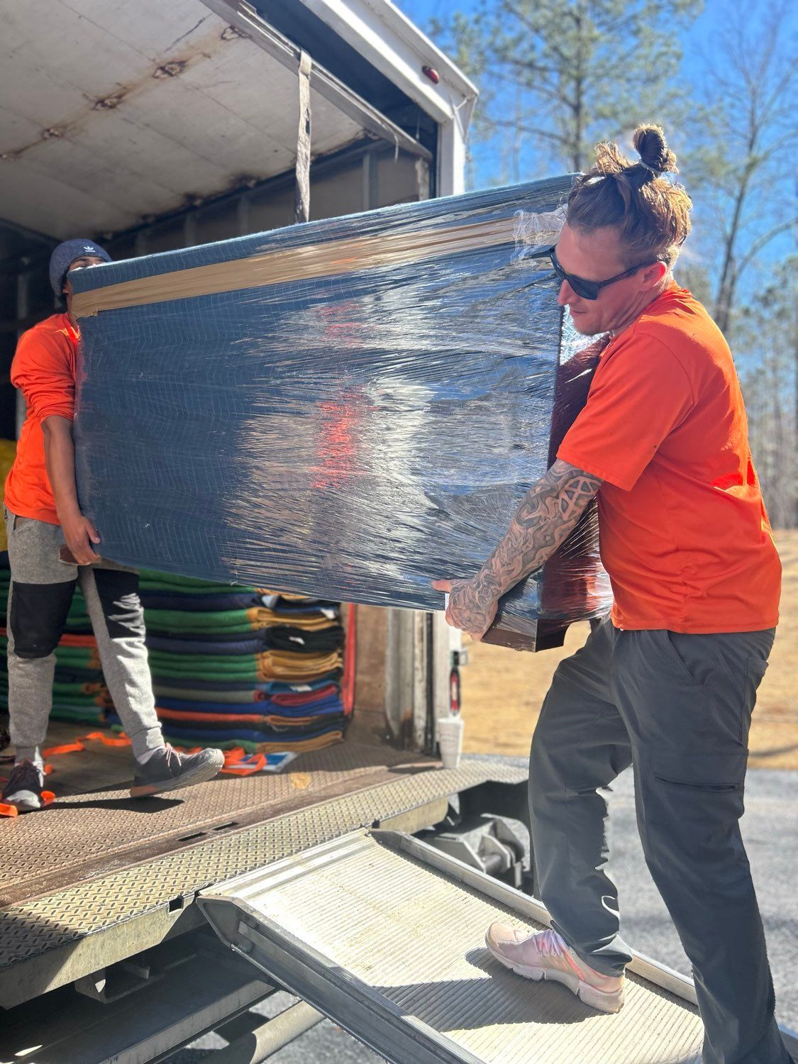Two workers in orange shirts unloading a wrapped box from a truck ramp