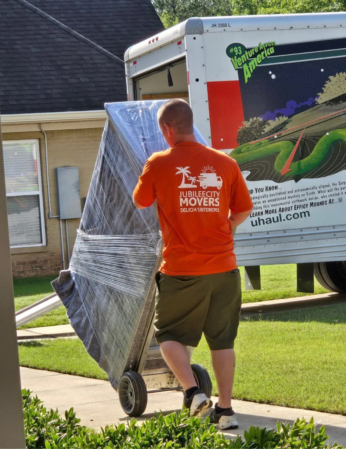 Worker in orange shirt and shorts pushing a wrapped appliance dolly beside a moving truck