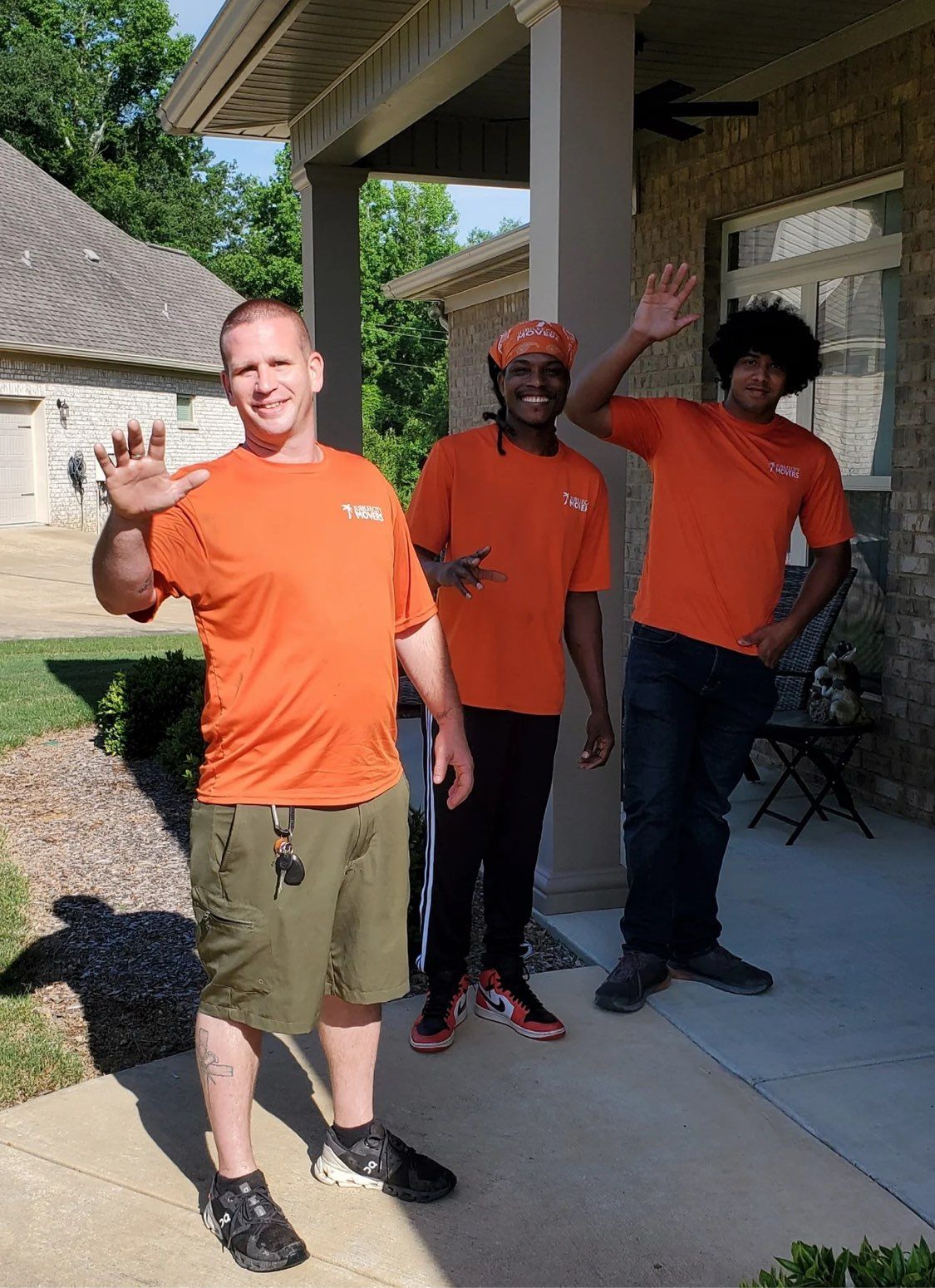 Three people in orange shirts waving on a front porch outside a house