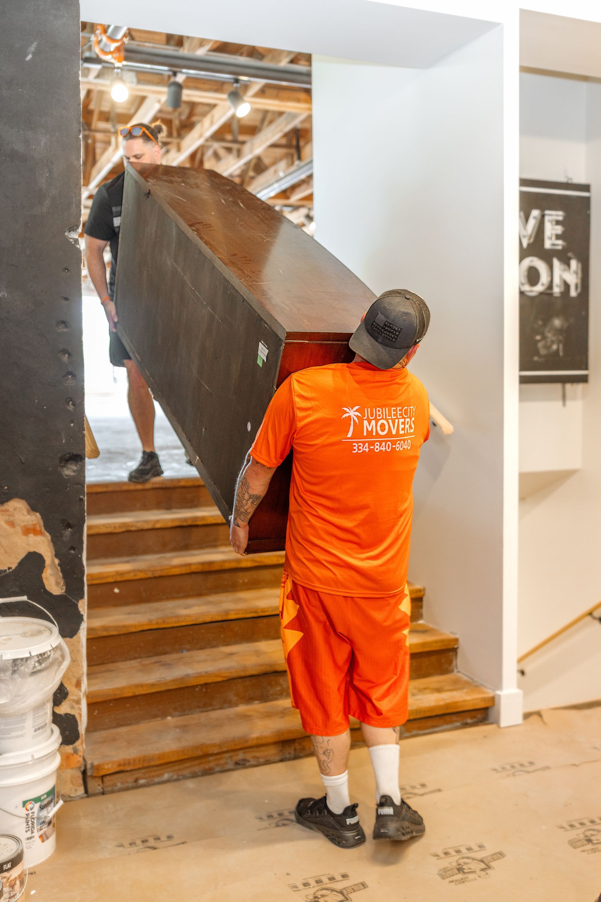 Worker in orange uniform carrying a large wooden beam up indoor stairs.