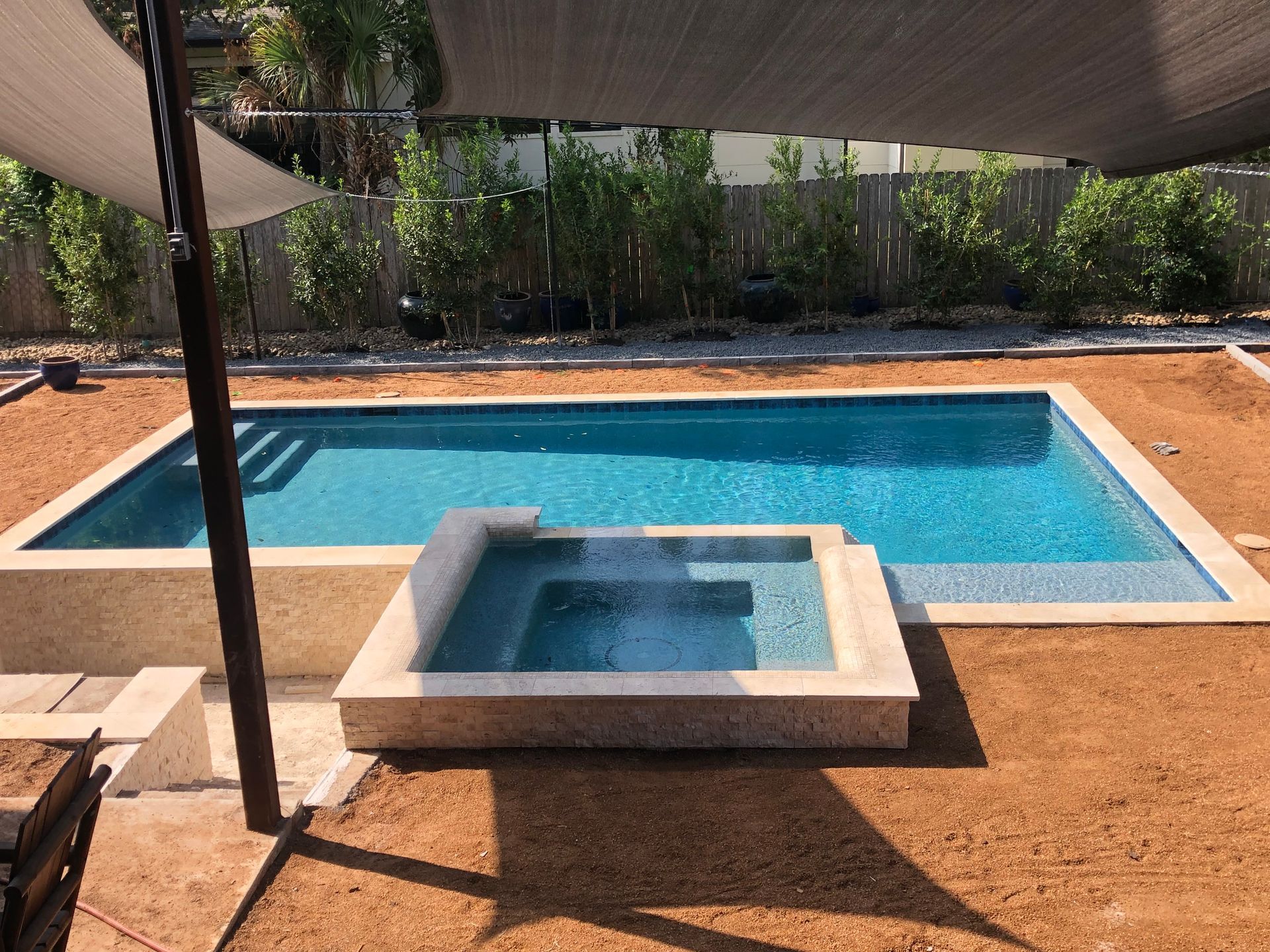 Pool with built-in spa, shade sails overhead, in a backyard with stone and brown ground cover.