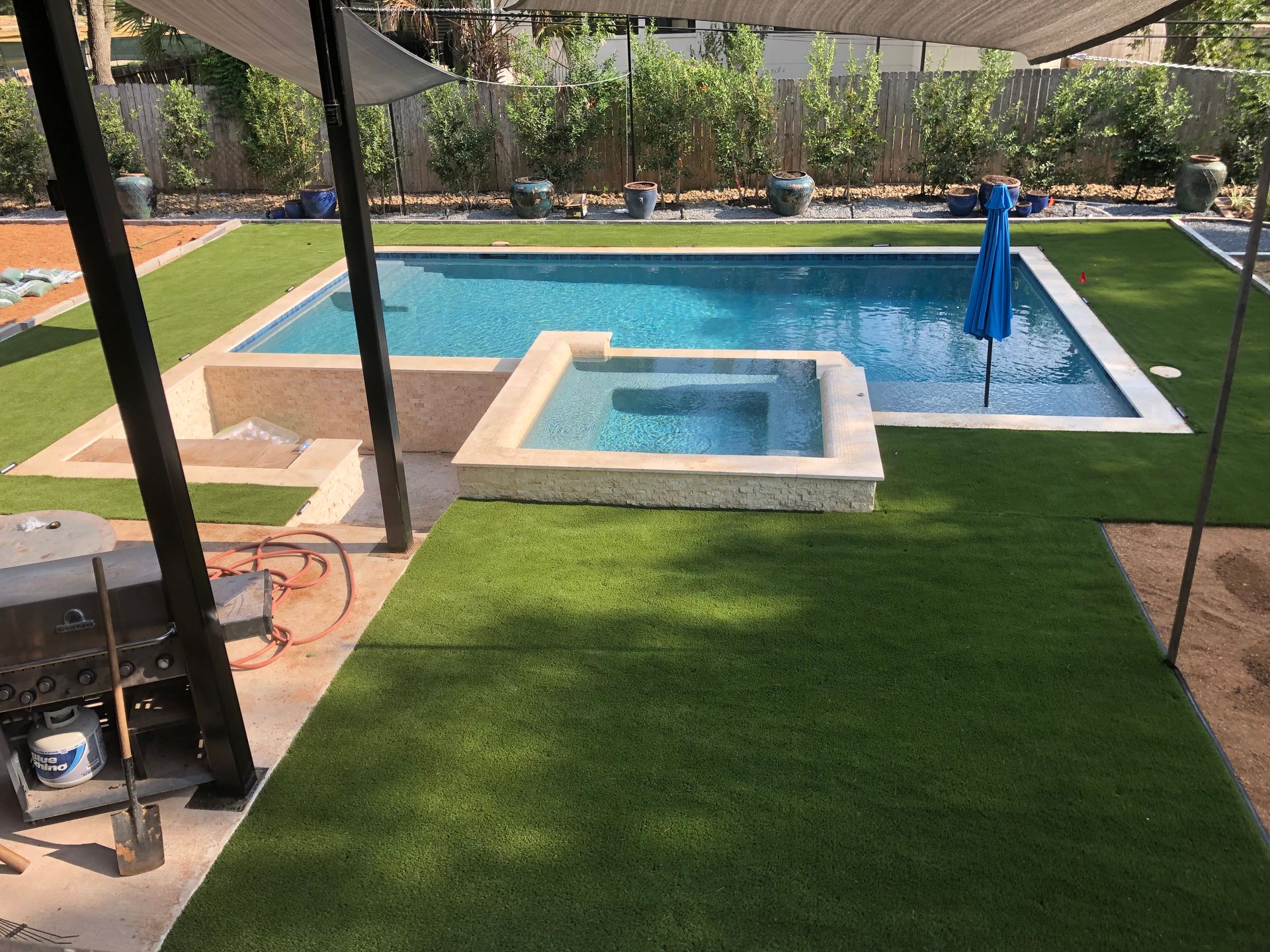 Backyard with rectangular pool and hot tub, surrounded by green turf. Blue umbrella in pool, patio cover overhead.