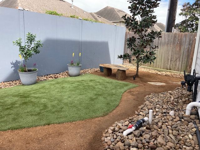 A small backyard with artificial grass, gravel path, and bench surrounded by a gray fence and plants.