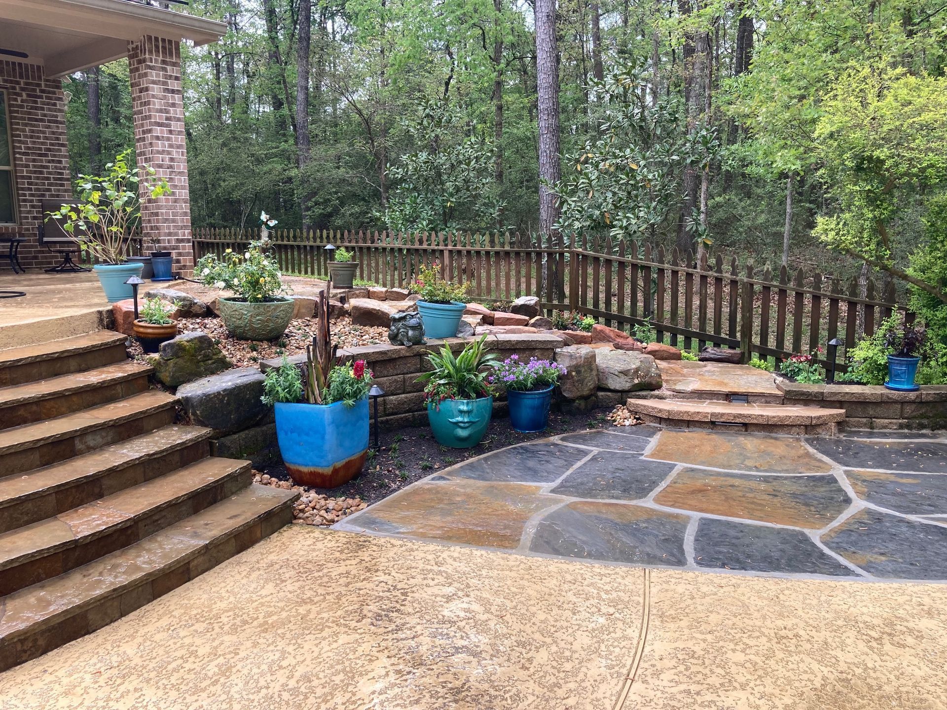 Patio with stone and concrete, tiered garden with blue pots, wooden fence, trees in background.