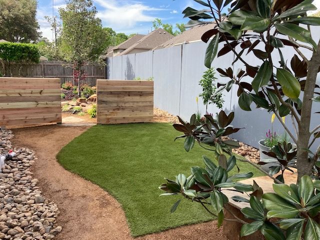 A backyard with a small artificial grass area, a gravel path, wooden fences, and a magnolia tree.