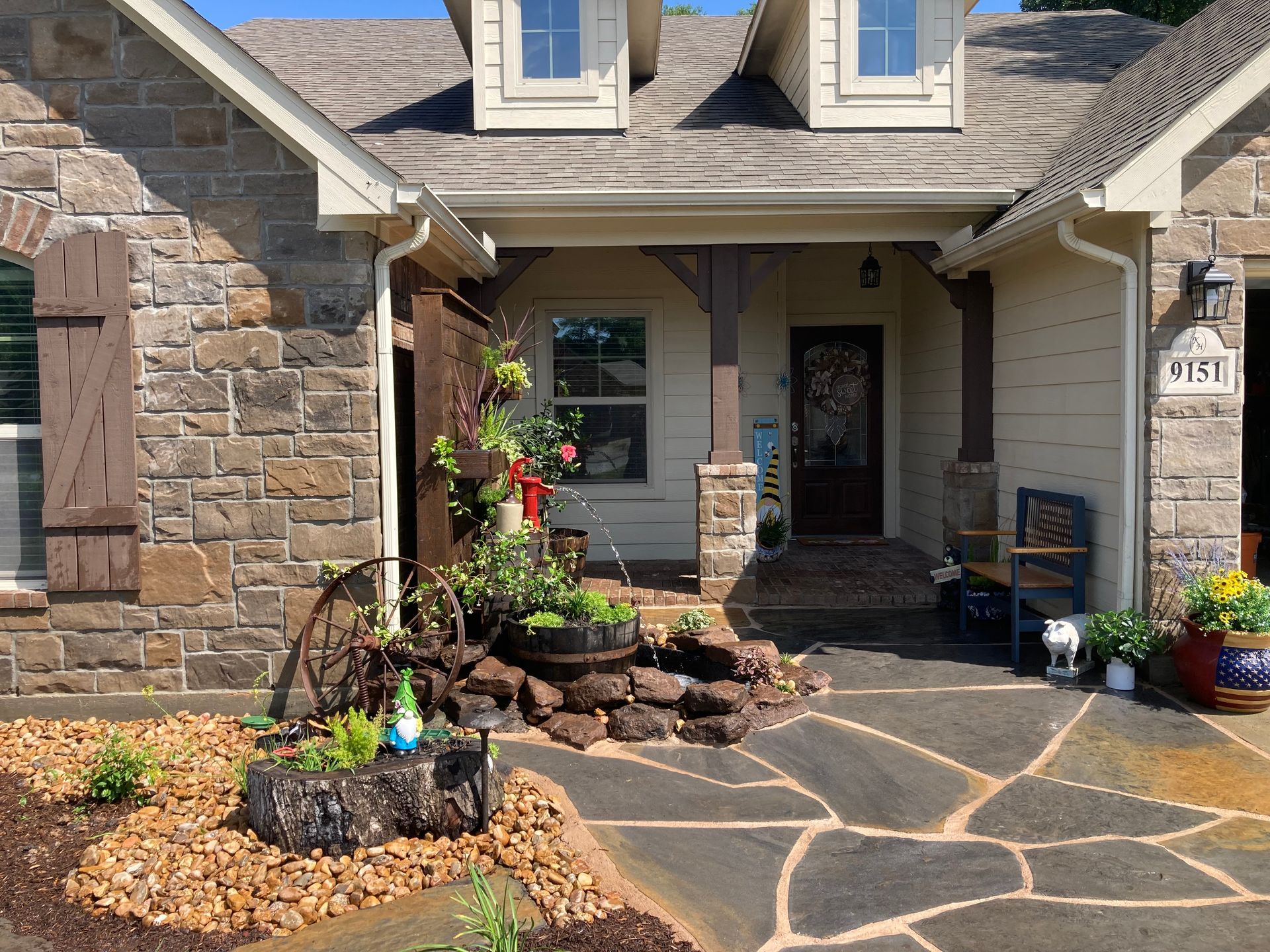 Stone-clad house with a walkway, front porch, and garden. Brown, gray, and green tones; flowers and foliage.