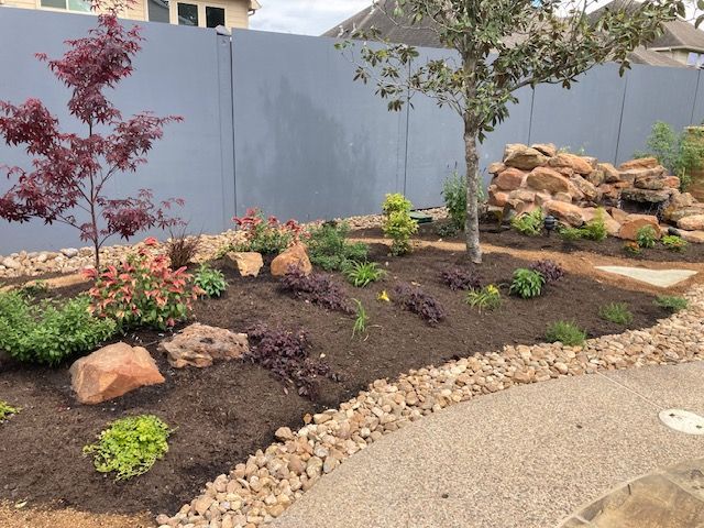 Landscaped garden bed with various plants and rocks, next to a gravel path and a gray fence.