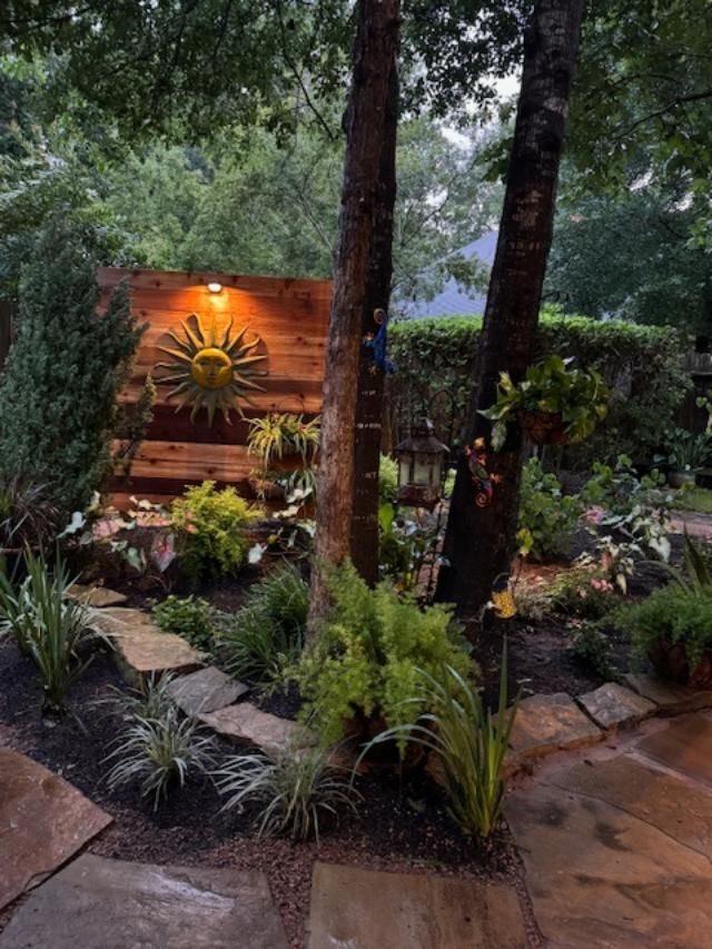A stone pathway leads through a lush garden with trees, plants, and a wooden backdrop with a sun decoration.